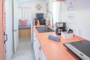 Una cocina compacta con mostrador, pileta y electrodomésticos. Se ve una cama con un almohadón rosa al fondo.