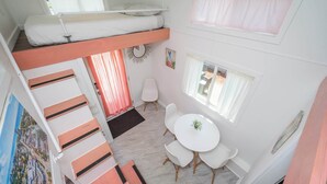 A room with a bunk bed, a dining table with chairs, a small white chair, a painting, and a window with curtains.