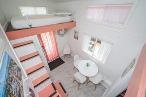 Une pièce avec un lit superposé, une table à manger avec des chaises, une petite chaise blanche, un tableau et une fenêtre avec des rideaux.