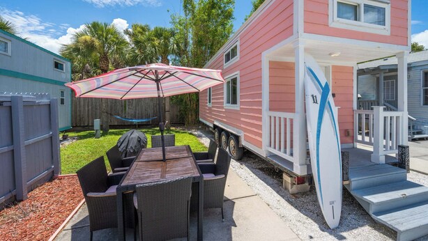 A pink mobile home with a patio area featuring a table and chairs, a striped umbrella, and a surfboard leaning against the house.