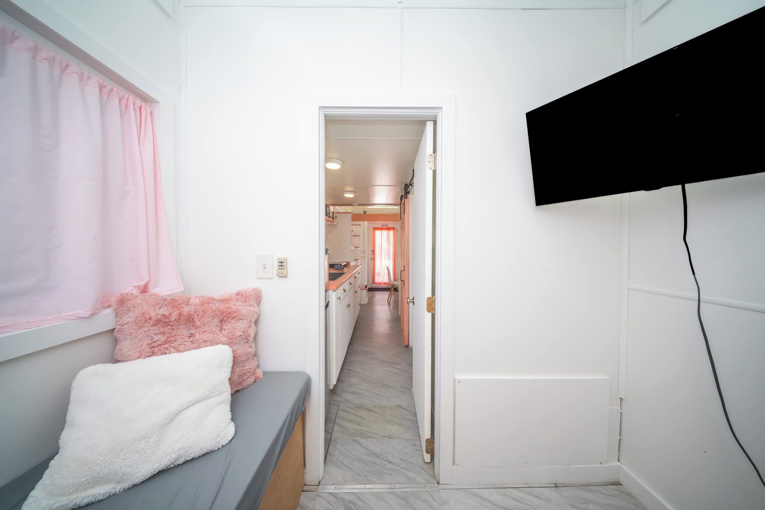 Um corredor estreito com uma cama à esquerda, uma televisão fixada na parede e uma porta que dá para uma cozinha.