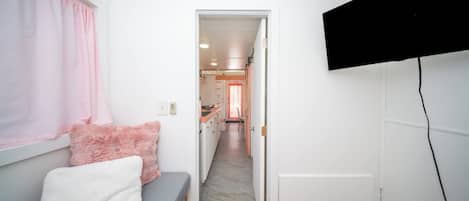 A narrow hallway with a bed on the left, a TV mounted on the wall, and a door leading to a kitchen area.