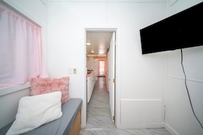 Un pasillo estrecho con una cama a la izquierda, un televisor montado en la pared y una puerta que da a una zona de cocina.