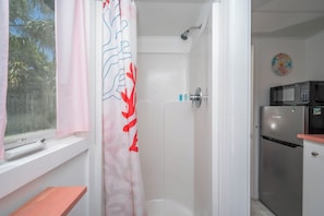 Een badkamer met een douche, toilet en een raam met gordijn. In de aangrenzende keukenruimte staat een magnetron en een koelkast.