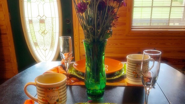 Una mesa de comedor con un jarrón verde de flores, un plato con la inscripción 'happy fall' y un vaso.