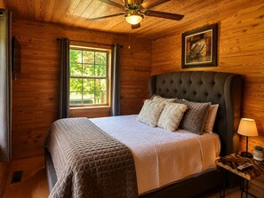 Un dormitorio con una cama grande, un ventilador de techo, una ventana con cortinas y un cuadro enmarcado en la pared.