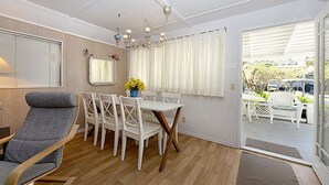 A dining area with a white table and chairs, a grey lounge chair, a chandelier, and a view through a glass door. - 334S: Quaint Catalina Cottage, Large Porch, BBQ, WIFI, Min Away from Beach - 334 (Avalon)