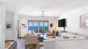 Un vaste salon doté d’une grande fenêtre offrant une vue sur la mer, d’un téléviseur à écran plat fixé au mur et d’un coin salon confortable avec un canapé et des fauteuils.
