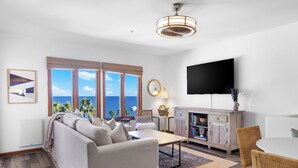 Un salon moderne avec un canapé, une table basse et un coin repas. Une grande fenêtre offre une vue sur la mer. Un téléviseur à écran plat est fixé au mur.