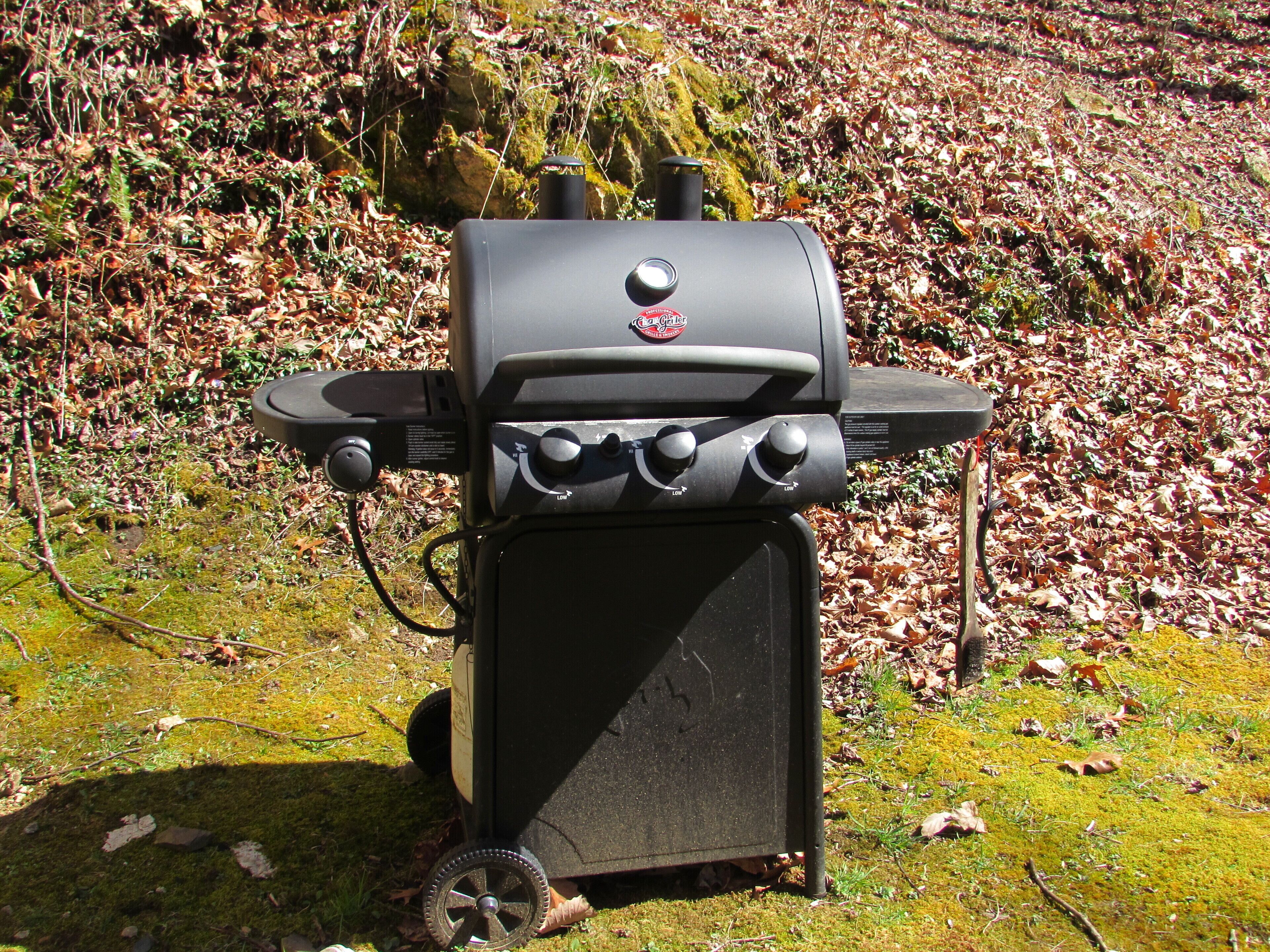 A portable grill with four burners and a control panel, set on a grassy area with fallen leaves.