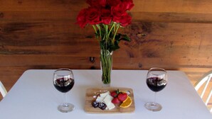 Une table sur laquelle se trouve une planche à découper en bois, avec du fromage, des fraises et une tranche d’orange, accompagnée de deux verres de vin rouge et d’un vase de roses rouges.