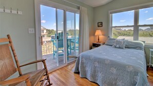 A bedroom with a large bed, a wooden rocking chair, and a view of the outdoors through a sliding glass door. - Discover ocean views at Moonlighting, a spacious 4-bedroom home in Topsail Beach! (Topsail Beach)