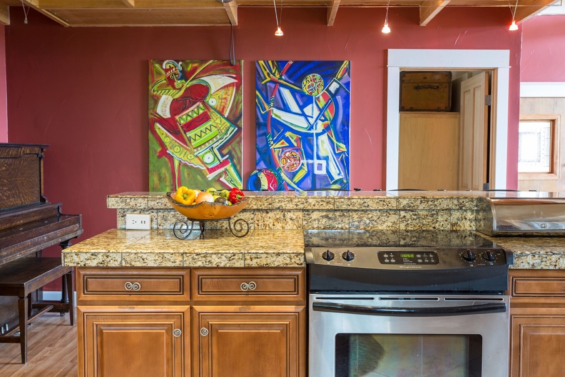 A kitchen with granite countertops, wooden cabinets, a stove, and two colorful abstract paintings on the wall.