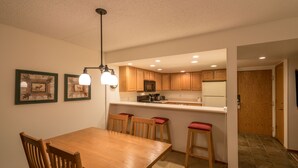 Un coin repas avec une table et des chaises en bois, une cuisine avec des meubles en bois et un plan de travail blanc, et un couloir menant à une porte.