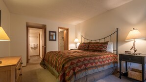 Une chambre à coucher comprenant un lit, des lampes de chevet, une commode en bois et une porte donnant sur une salle de bain.