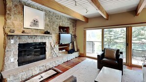 A stone fireplace with a TV above it, a leather sofa, and a large window with a view of trees. - 🌲Timberline D2D Deluxe 2-Bedroom ~Hot Tub~Pool~WiFi~Ski-in/out~Local Shuttle🌲 (Snowmass Village)