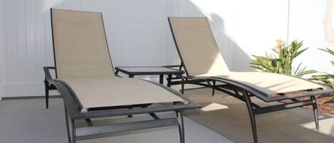 Two lounge chairs with metal frames and beige cushions, positioned on a concrete patio with a white wall in the background.