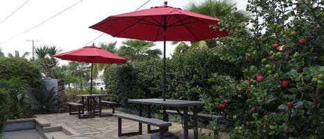 Een terras buiten met stenen vloer, picknicktafels en rode parasols.
