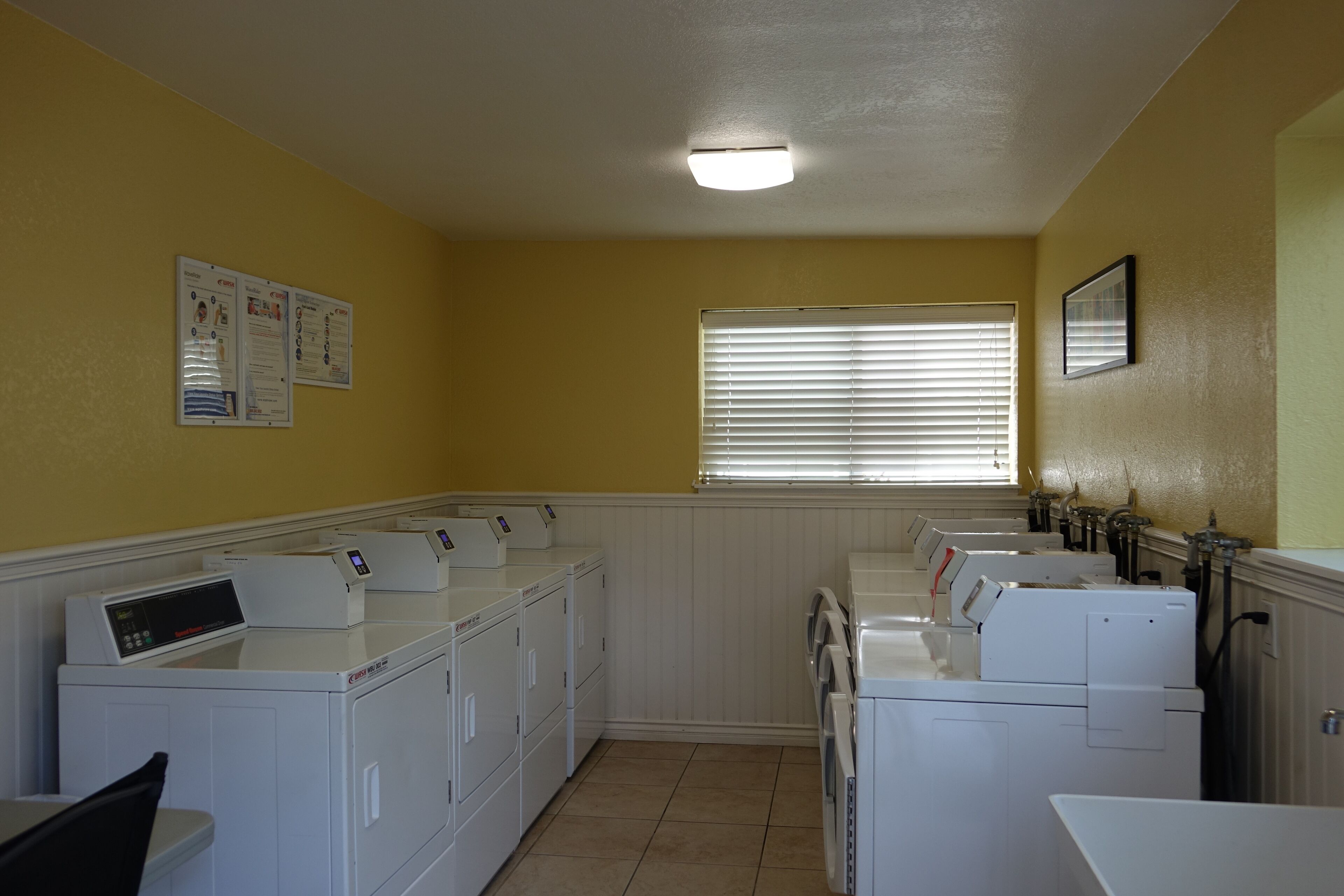 A laundry room with multiple washing machines and dryers, a window with blinds, and a bulletin board on the wall.
