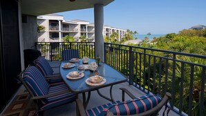 A balcony with a table set for dining, surrounded by striped cushioned chairs, overlooking a resort with multiple buildings and a clear view of the ocean. - Carolina to Keys Ocean view dream Monthly pricing Best pool on the Island (Key West)
