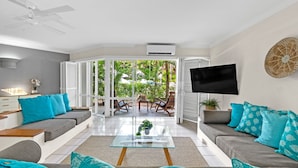 Una sala de estar moderna con sofá, mesa de centro y televisor. Hay un ventanal corredero que da a un área exterior con patio y sillas.