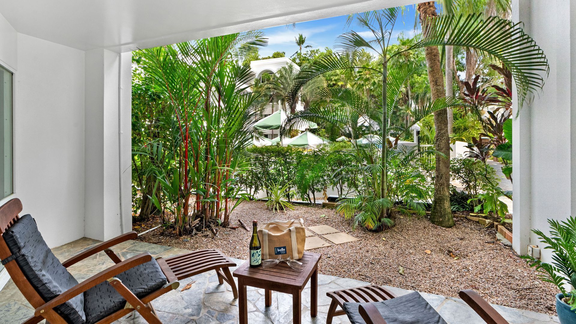 A covered outdoor seating area with wooden chairs and a table, surrounded by lush tropical plants and a view of a white building in the background.