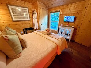 A wooden cabin room with a bed, a television, and a window with a view of greenery. - Apple Blossom Cottage Of Gettysburg.  Hot Tub is an Add on Fee (Orrtanna)
