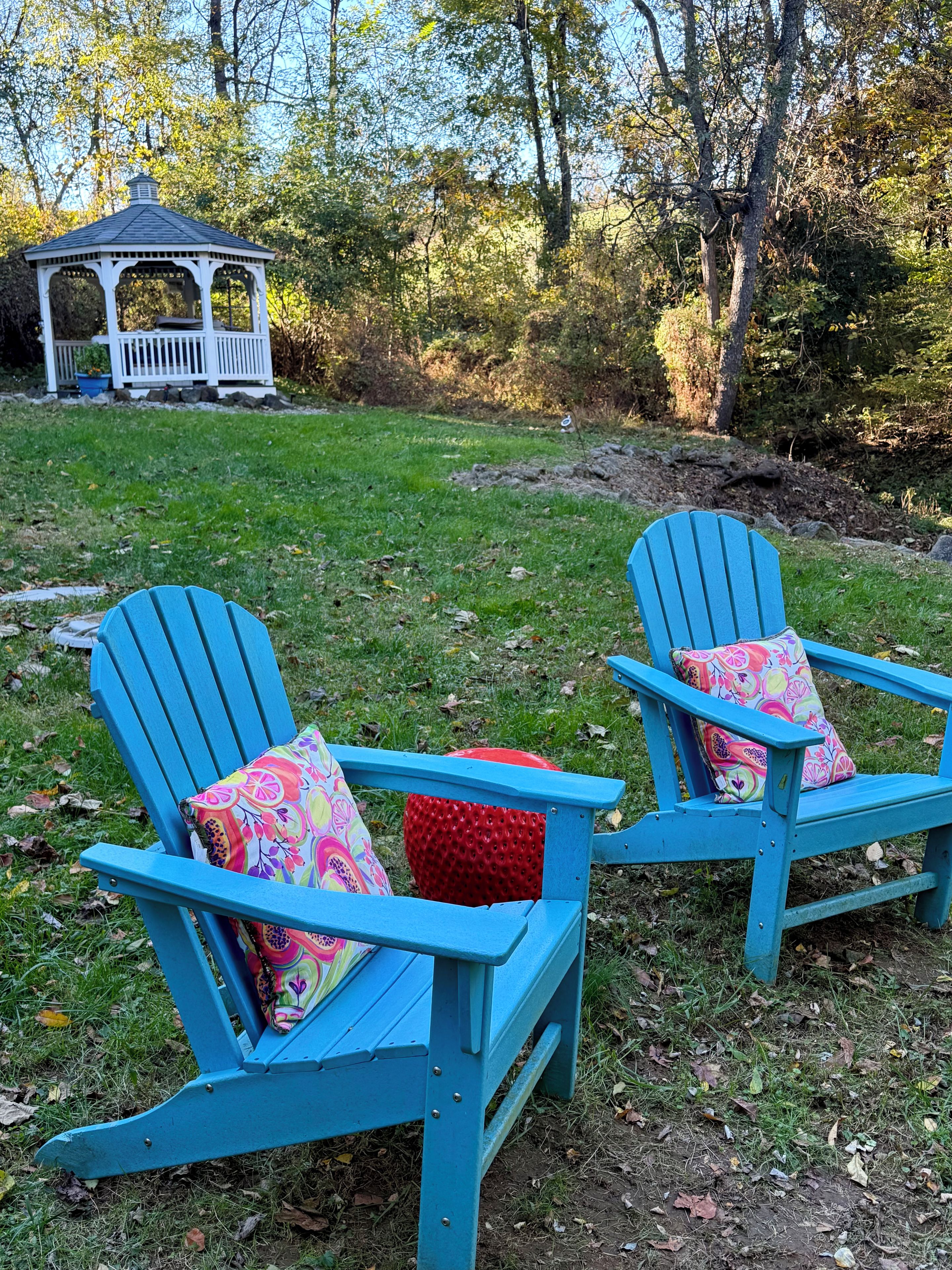 Deux fauteuils Adirondack avec des coussins colorés, placés dans un espace verdoyant, avec un gazebo en arrière-plan.