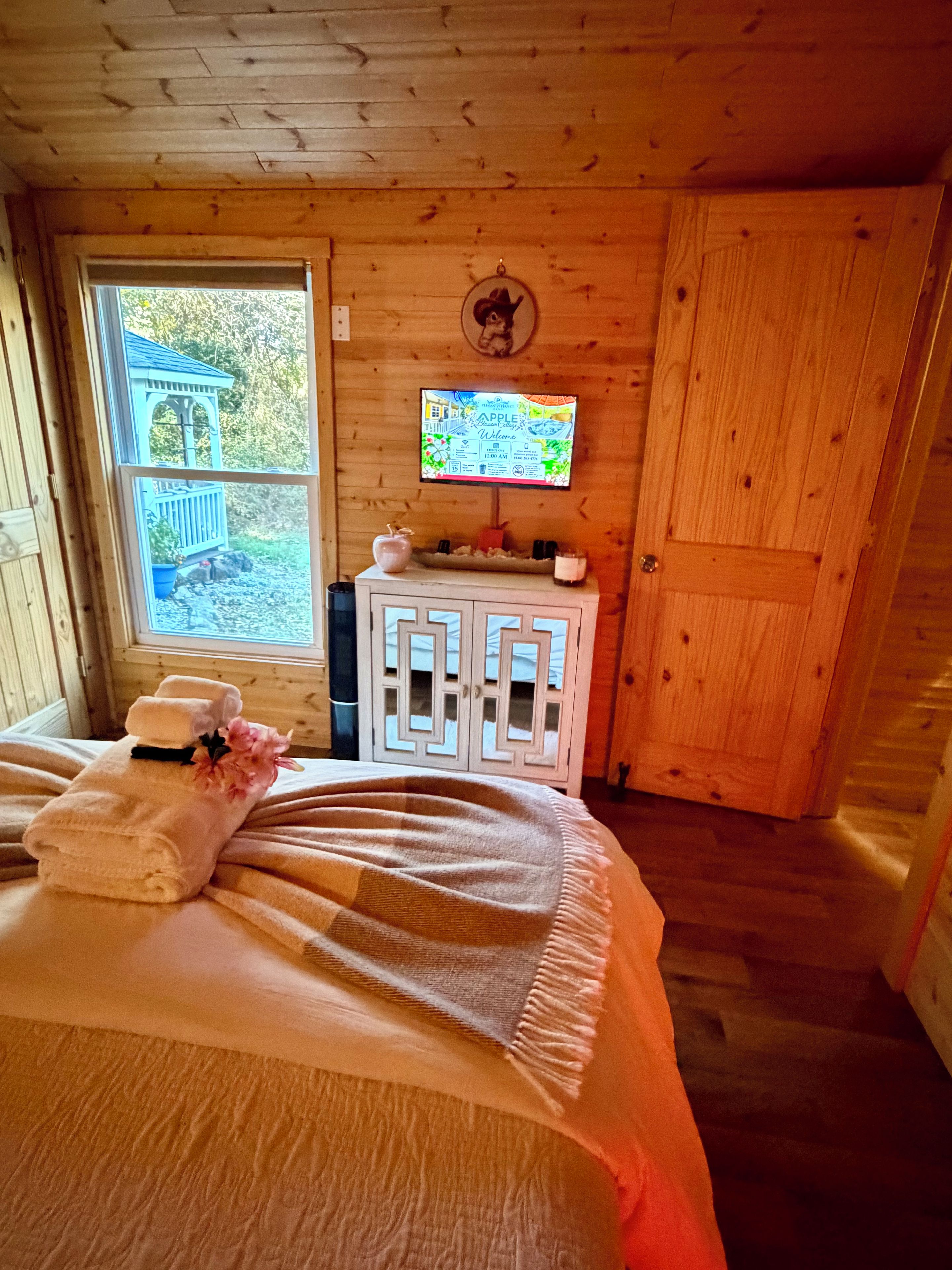A wooden cabin room with a bed, a television on the wall, and a window with a view of a wooden deck.