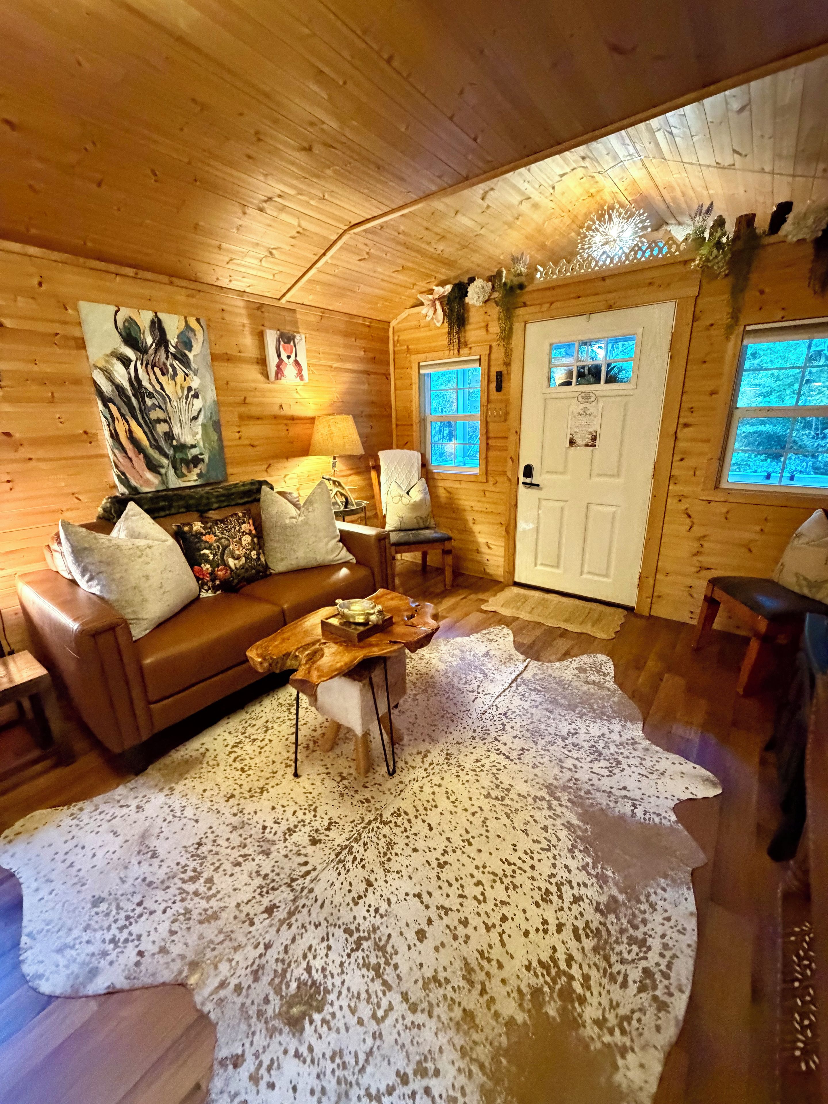 A cozy cabin interior with a wooden ceiling, a brown leather sofa, a coffee table, and a large white rug.