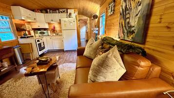 A cozy interior with a brown leather sofa, a wooden dining table, and a kitchen area with white cabinets and a refrigerator.