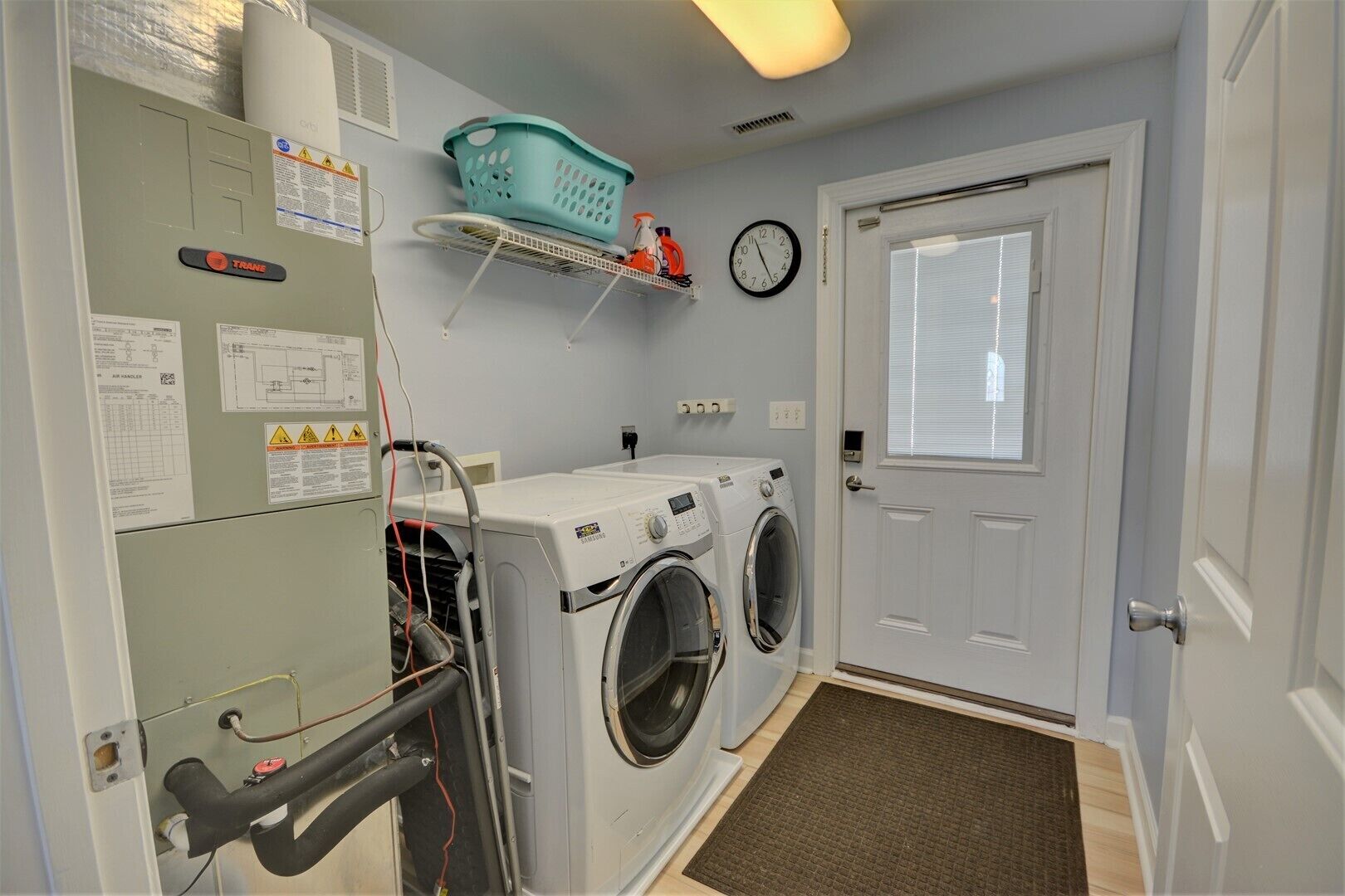 A laundry room with two front-loading washers and dryers, a wall-mounted clock, and a small shelf with a laundry basket and cleaning supplies.