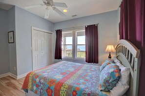 A bedroom with a bed, a ceiling fan, a window with curtains, and a view of the sea.