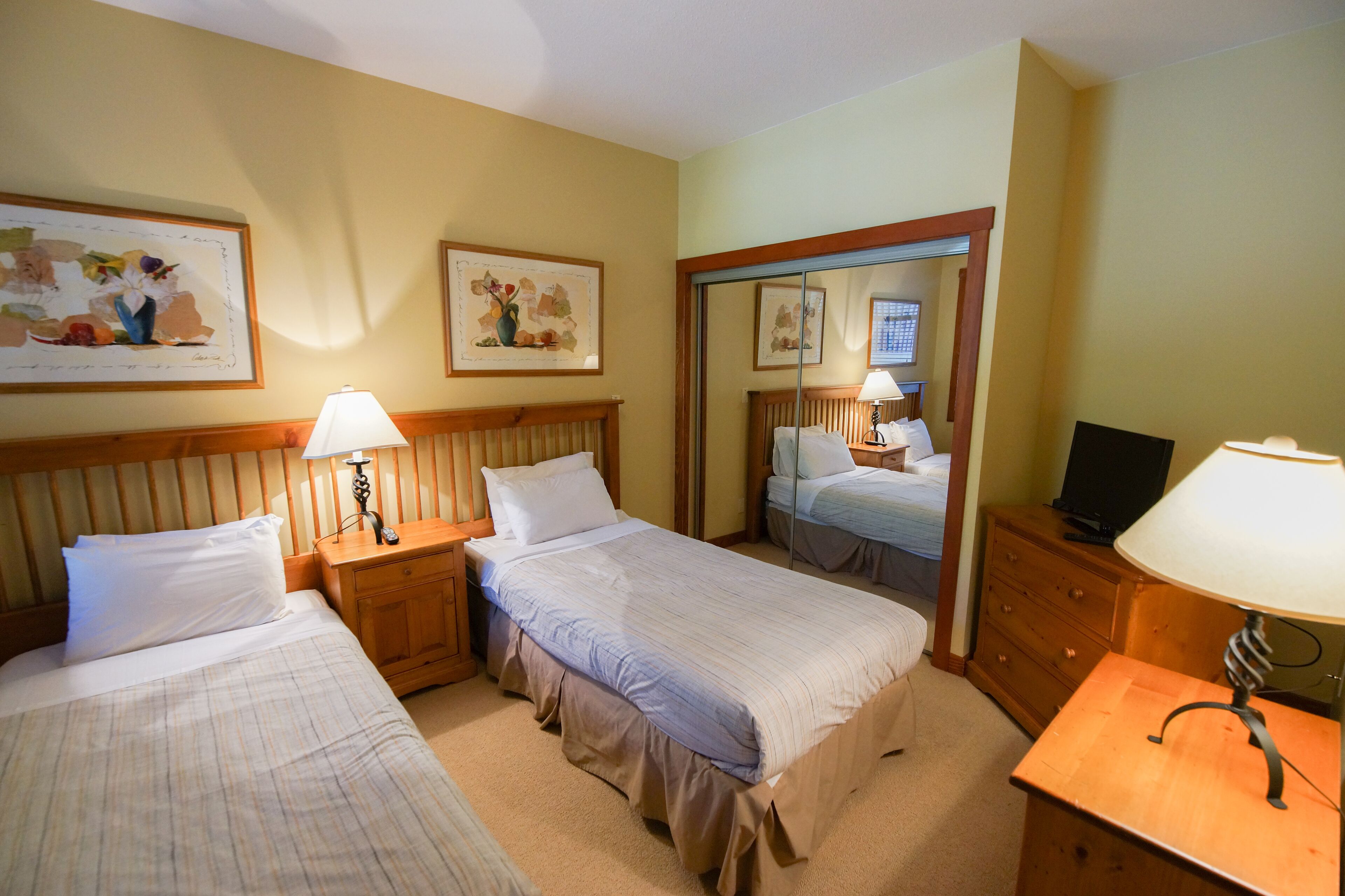 Habitación de hotel con dos camas, cabeceros de madera, mesitas de noche con lámparas, cómoda y cuadros en las paredes.