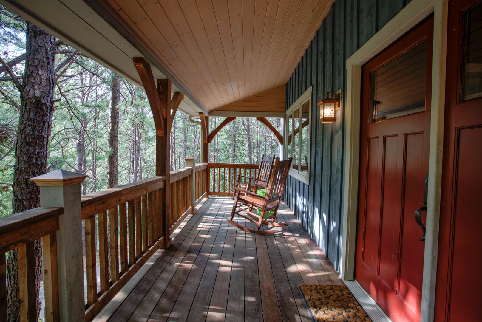 Un portico in legno con una sedia a dondolo, una porta rossa e vista sugli alberi.
