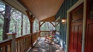 A wooden porch with a rocking chair, a red door, and a view of trees.