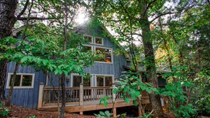 A rustic cabin with a wooden deck, surrounded by trees and foliage.