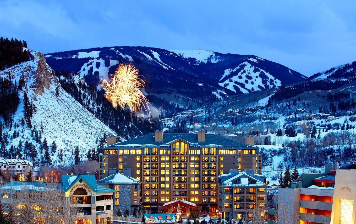 A hotel with multiple floors, a prominent building in the center, and snow-covered mountains in the background.