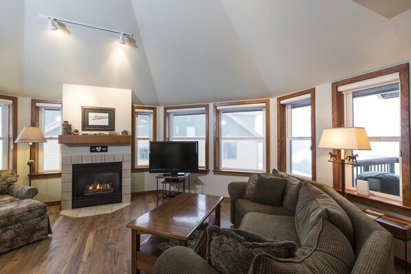 A cozy living room with a fireplace, a television, and a large window overlooking a balcony.