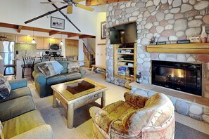 Una sala de estar con chimenea de piedra, un televisor montado encima y una cocina al fondo.