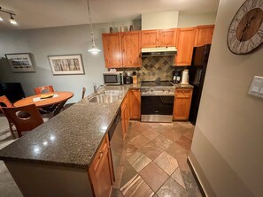 A kitchen with granite countertops, wooden cabinets, a stainless steel sink, a microwave, a stove, and a clock on the wall.