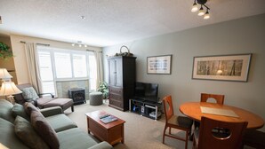 A living room with a sofa, a coffee table, a TV, a cabinet, and a dining table with chairs. - Glacier Lodge - Deluxe Ski in/Ski out (Whistler)