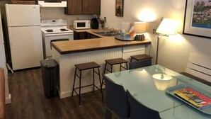 A compact kitchen with a refrigerator, stove, microwave, and a small sink. There are wooden cabinets, a bar stool, and a table with a magazine. - Iconic Sun Valley - Parkside Village - Sleeps 7 very comfortably  (Ketchum)