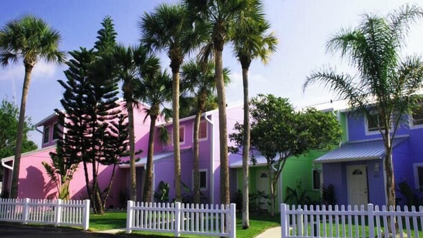A row of beach cottages with a white picket fence, palm trees, and a clear sky. - Spacious, Pet Friendly, Townhome short walk to the beach with backyard (New Smyrna Beach)