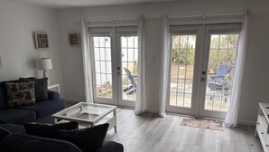 A living room with a dark blue sofa, a glass coffee table, and a white side table. There are two sliding glass doors leading to an outdoor area with a patio and chairs. - Spacious, Pet Friendly, Townhome short walk to the beach with backyard (New Smyrna Beach)