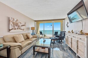 Un salón amplio con un balcón con vista a la playa, un televisor de pantalla plana montado en la pared y un sofá de cuero con cojines estampados.