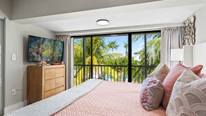 A bedroom with a large bed, a wooden dresser, a television, and a balcony with a view of palm trees. - NEW! Casa Palma - NO HIDDEN FEES! Golf Cart, 2 King, Pool, Beach, Tennis, Kayaks (Key Largo)