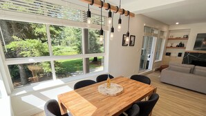 Een moderne eethoek met een houten tafel en stoelen, grote ramen met uitzicht op groen en een keuken op de achtergrond.