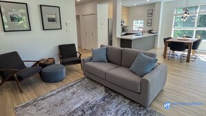 Een moderne woonkamer met een grijze bank, twee fauteuils, een salontafel en een vloerkleed. Een keuken met een eettafel en stoelen is zichtbaar door een open doorgang.