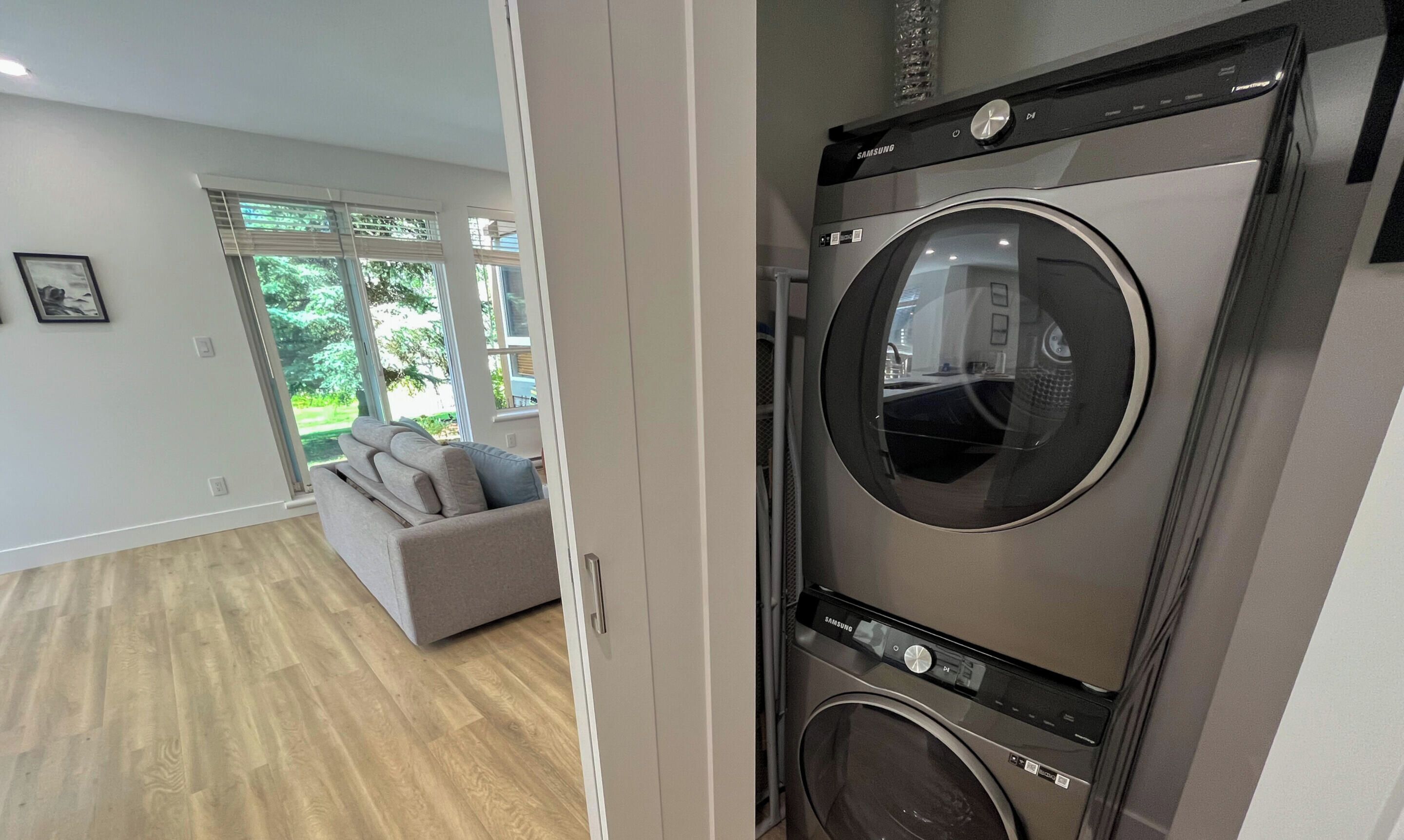 Un lavadero moderno con lavadora y secadora Samsung, un sofá gris en la zona de estar contigua, y ventanas grandes con vistas al exterior.
